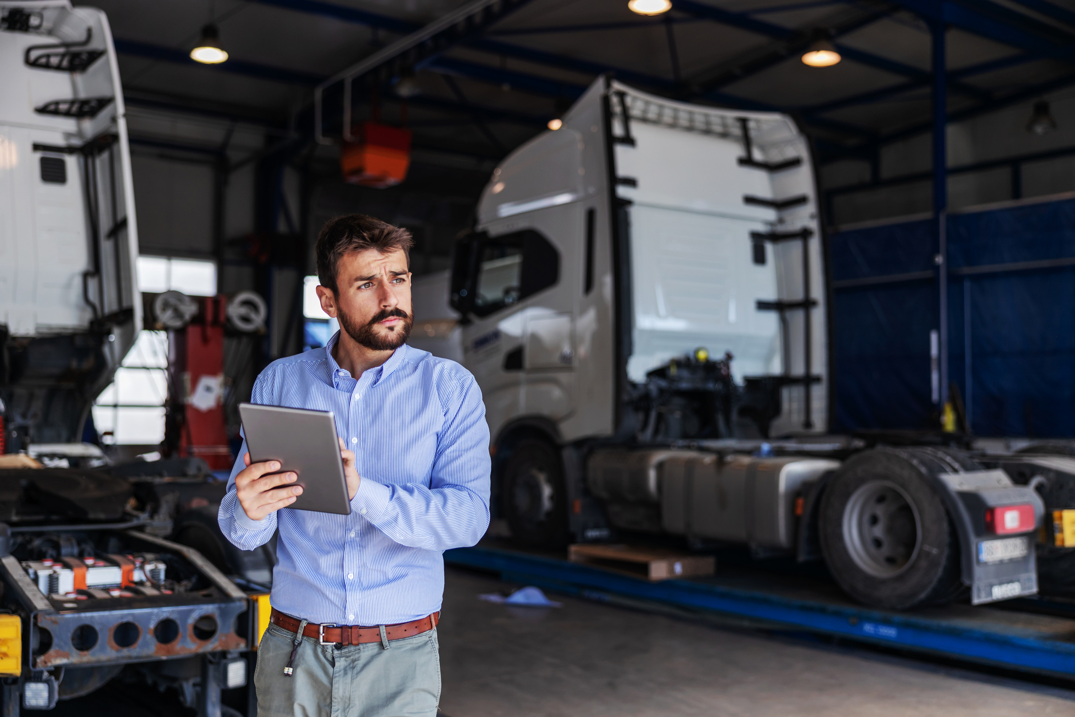 Young serious bearded CEO standing in garage of shipping firm and using tablet. In background are trucks