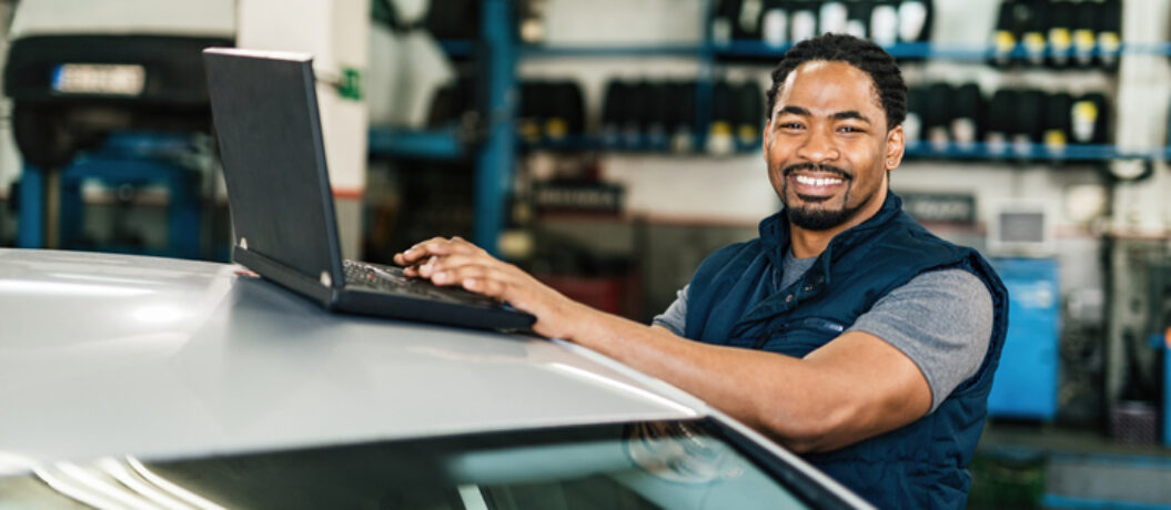 Happy black auto mechanic working on a computer and looking at camera in repair shop.