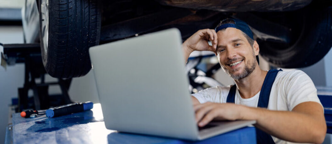 Happy car mechanic using laptop while working at auto repair shop and looking at camera.