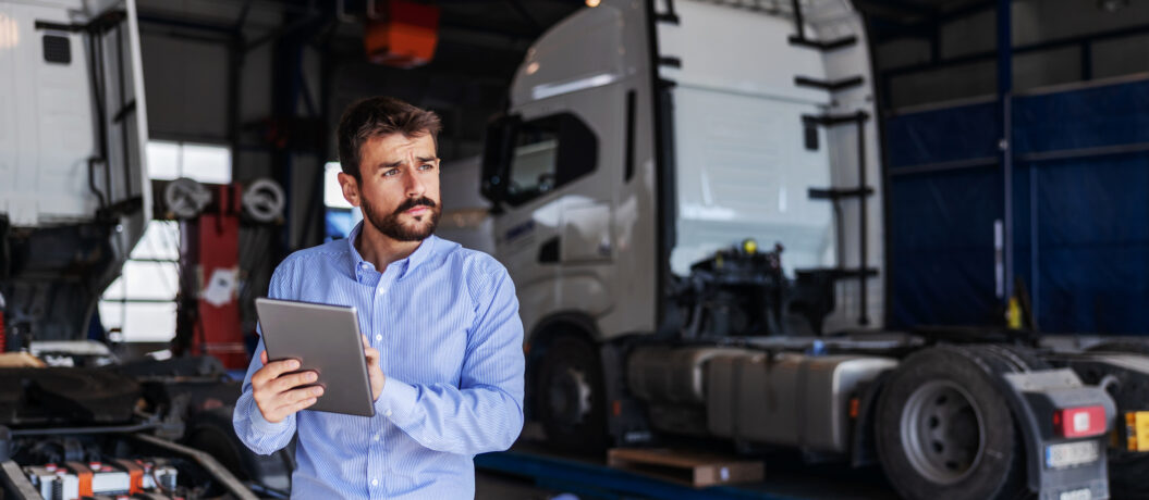 Young serious bearded CEO standing in garage of shipping firm and using tablet. In background are trucks.