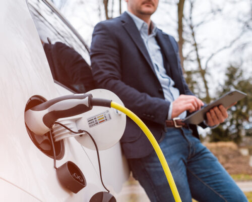 Business man standing near charging electric car or EV car and using tablet in the street at the sunlight.