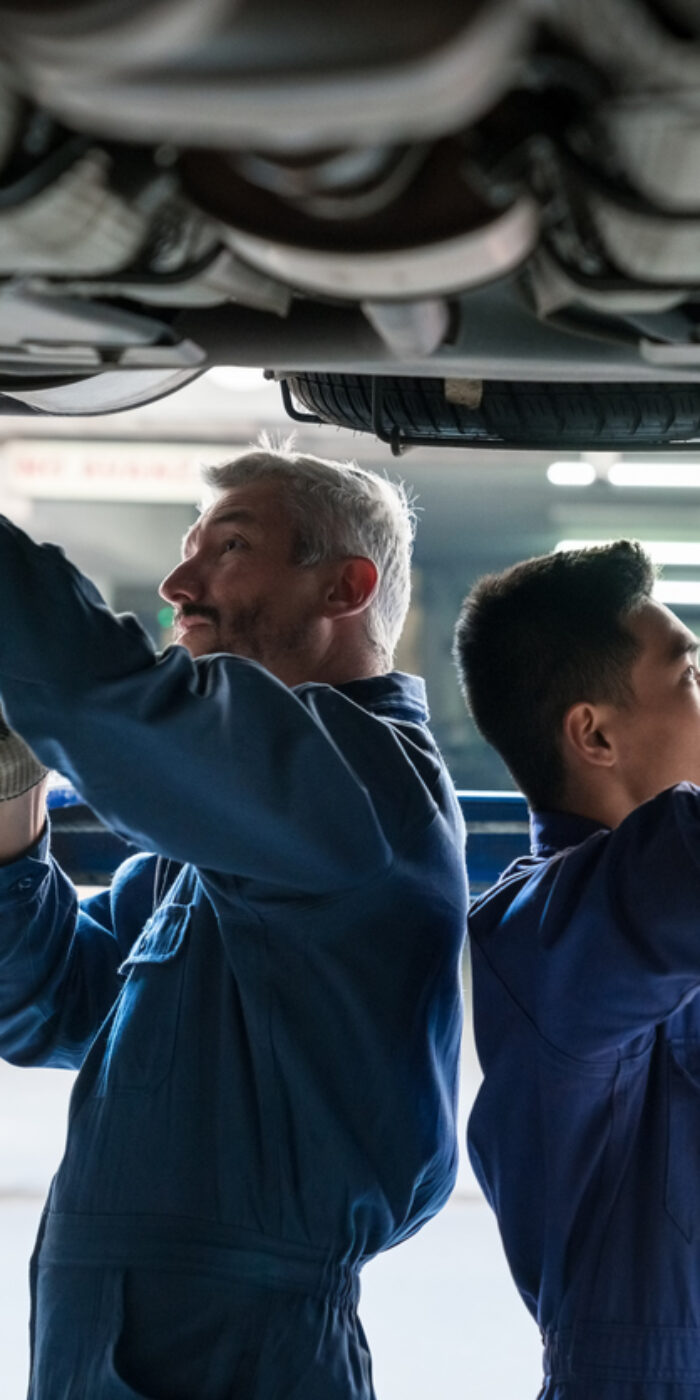 Side view of mature auto mechanic and young apprentice examining car in the auto repair shop, checking wheels.