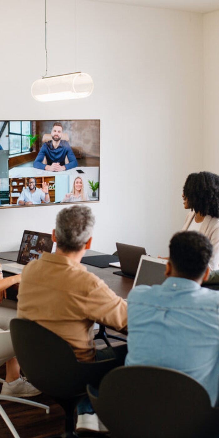 In a well-lit meeting room, a team actively engages with remote colleagues via a large screen, illustrating the modern hybrid work model where technology bridges the gap between in-office and remote team members.