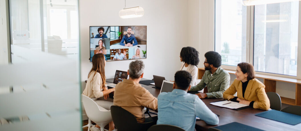 In a well-lit meeting room, a team actively engages with remote colleagues via a large screen, illustrating the modern hybrid work model where technology bridges the gap between in-office and remote team members.