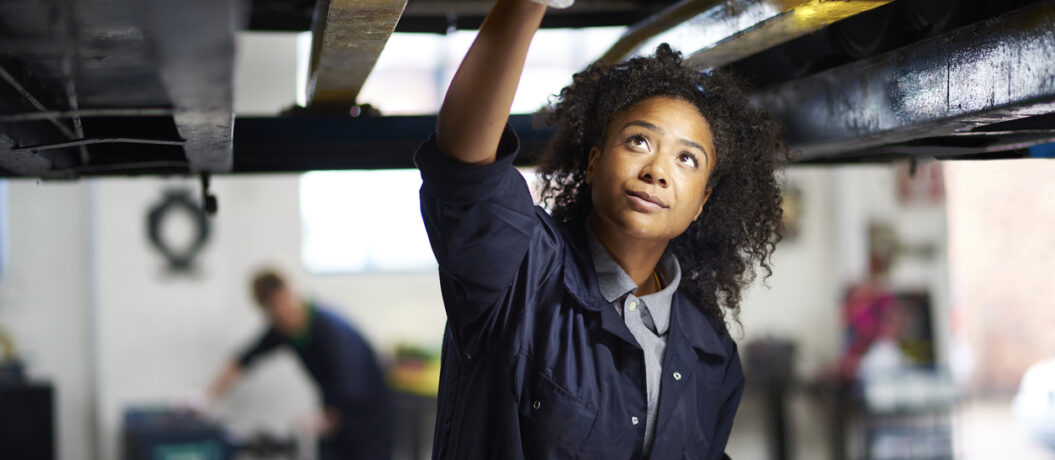 A young female adult mechanic is standing under a car in a garage. She is standing proudly and looking to camera
