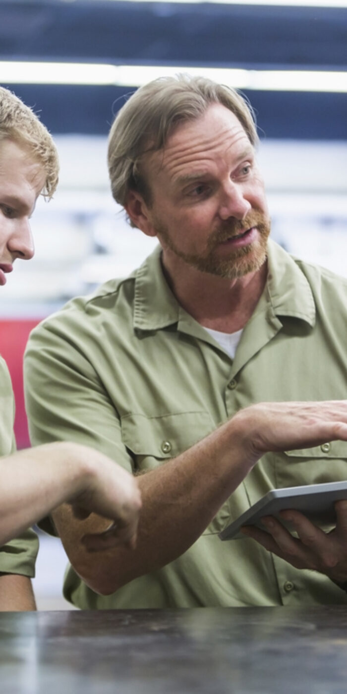 An instructor with three multi-ethnic adult students in vocational school, learning a trade. He is sitting with them at a table, holding a digital tablet, talking and looking at a young mixed race man sitting next to him.